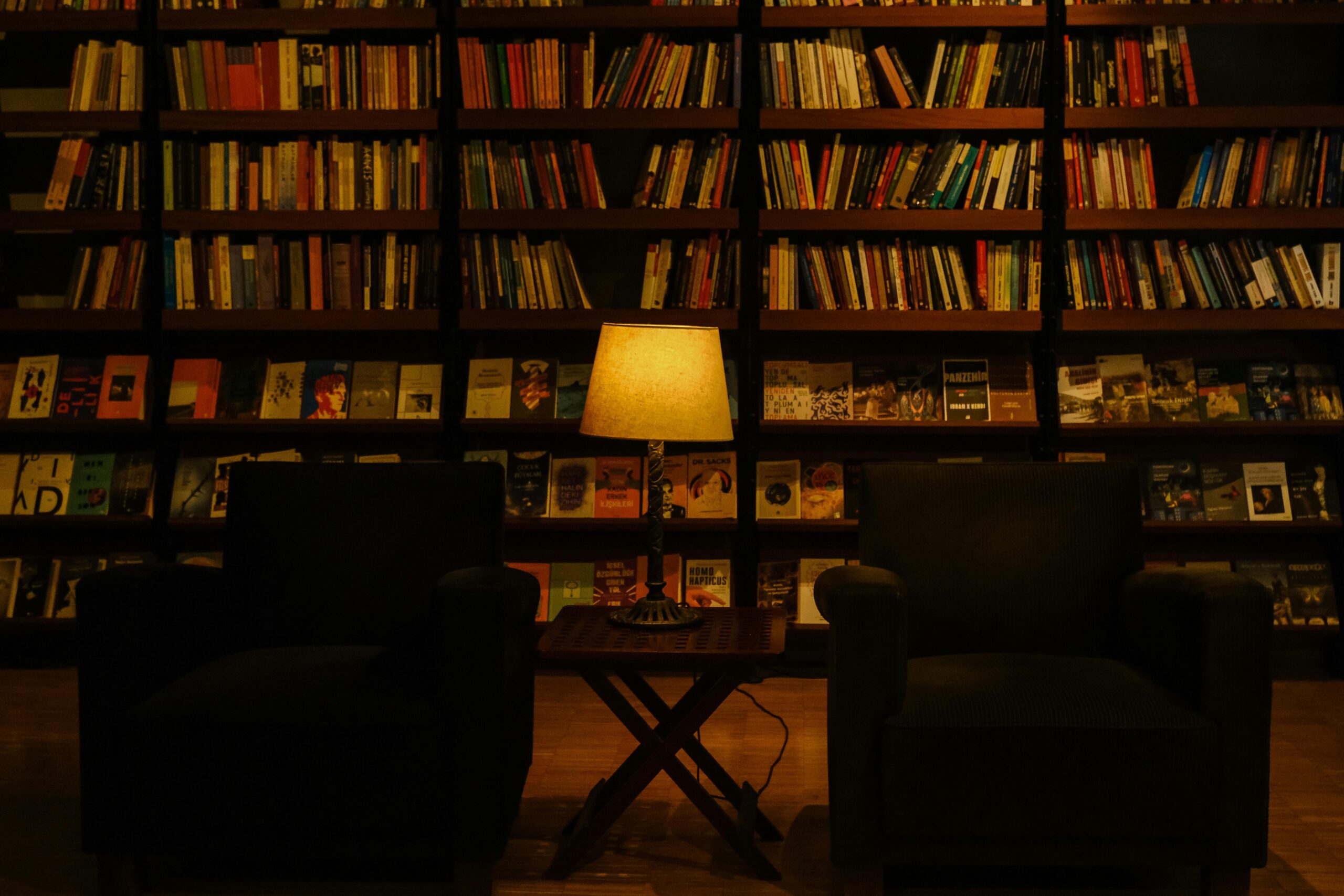 Dimly lit library setting featuring armchairs and a warm lamp, surrounded by bookshelves.